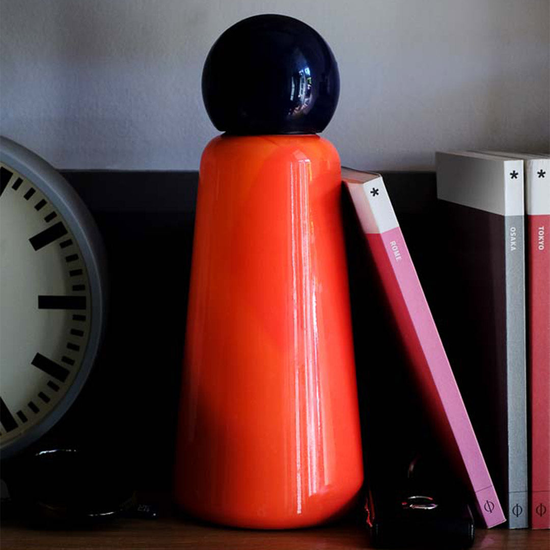 Lund London Coral & Indigo Skittle Water Bottle on a dark wooden shelf next to a clock on the left and books on the right 