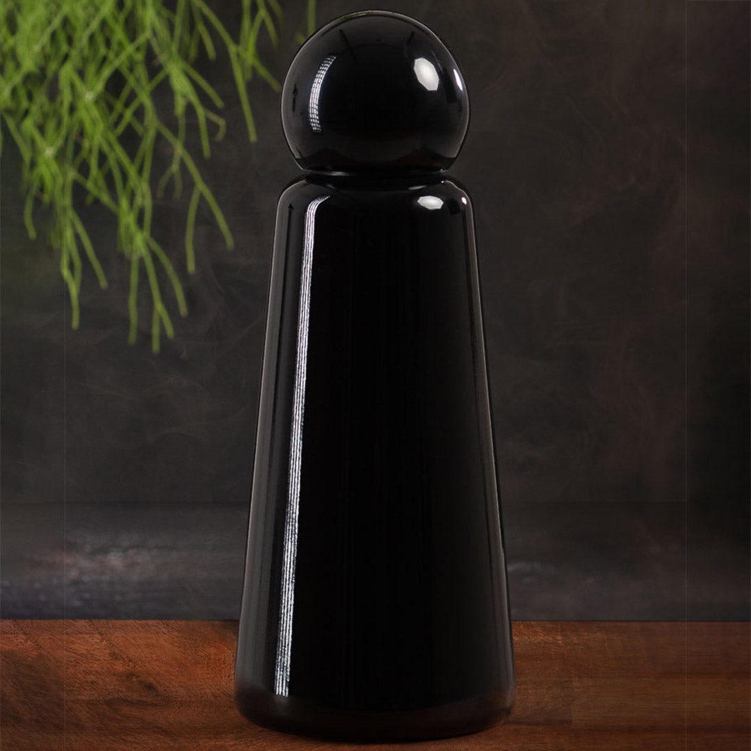 Skittle Bottle Original Midnight Black sat on a wooden table with a dark grey background and a plant dropping down from the top left side